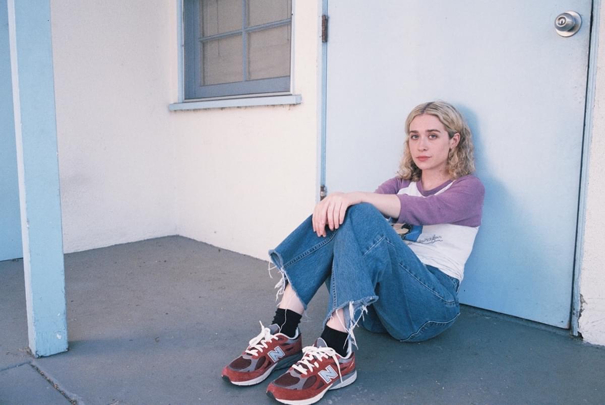 Blondshell sitting against blue door jeans red trainers