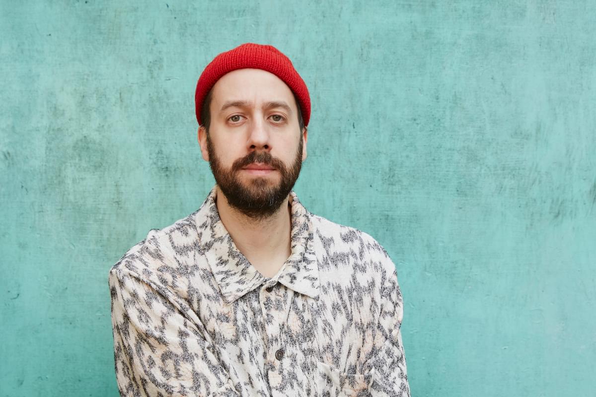 Gold Panda against a blue-green backdrop wearing a red beanie