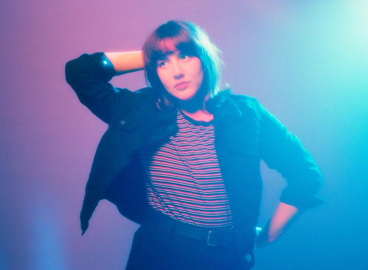 Caitlin Rose with one hand on her hip and the other behind her head, in blue and purple lighting